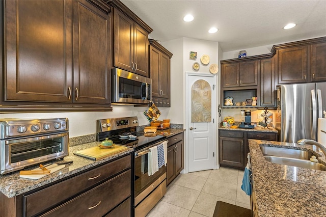 kitchen with dark brown cabinetry, sink, dark stone counters, light tile patterned floors, and appliances with stainless steel finishes