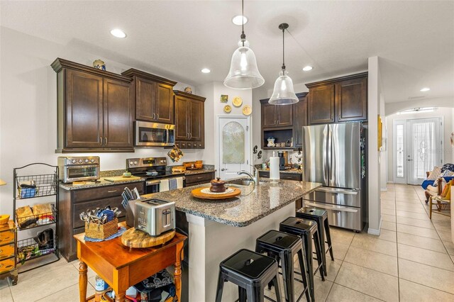 kitchen featuring dark stone counters, hanging light fixtures, an island with sink, appliances with stainless steel finishes, and dark brown cabinets