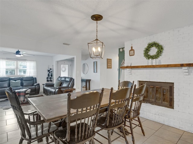 dining space featuring light tile patterned floors, visible vents, a brick fireplace, a textured ceiling, and ceiling fan with notable chandelier