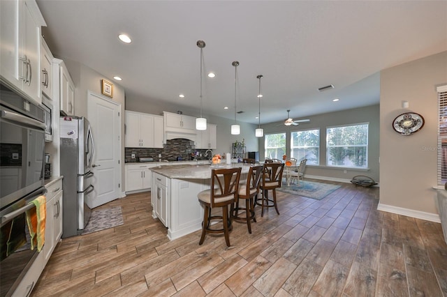 kitchen with pendant lighting, a kitchen island with sink, white cabinetry, wood-type flooring, and stainless steel refrigerator