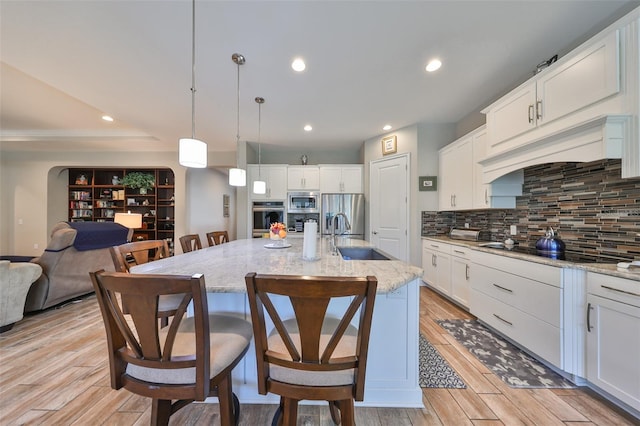 kitchen with light wood-type flooring, a kitchen island with sink, sink, white cabinetry, and hanging light fixtures