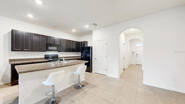 kitchen featuring a kitchen island with sink, black appliances, light tile patterned floors, a kitchen bar, and dark brown cabinets