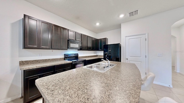 kitchen with sink, light tile patterned floors, black appliances, dark brown cabinetry, and a kitchen island with sink