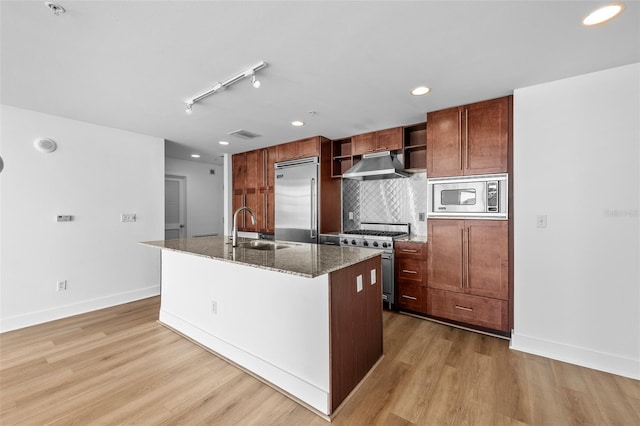 kitchen with dark stone counters, sink, built in appliances, an island with sink, and light hardwood / wood-style floors