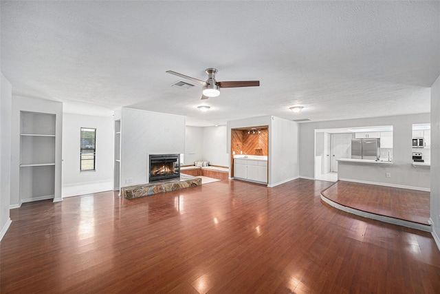 unfurnished living room featuring a stone fireplace, a textured ceiling, hardwood / wood-style flooring, built in features, and ceiling fan