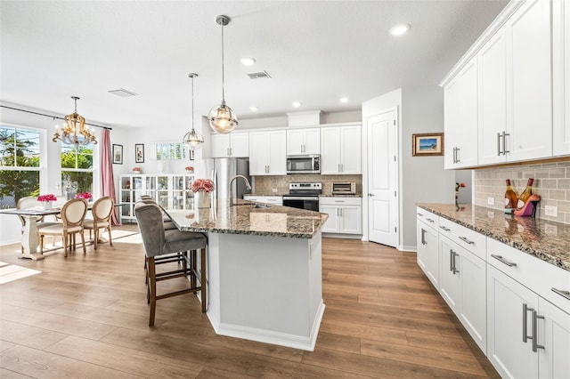 kitchen with stainless steel appliances, hardwood / wood-style flooring, dark stone countertops, white cabinetry, and an island with sink