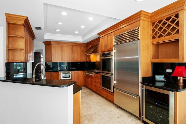 kitchen with kitchen peninsula, appliances with stainless steel finishes, a tray ceiling, wall chimney range hood, and wine cooler