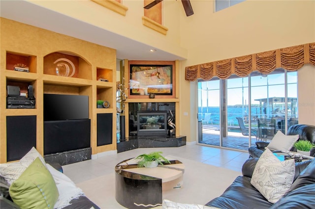 tiled living room with ceiling fan, a water view, built in features, a premium fireplace, and a high ceiling