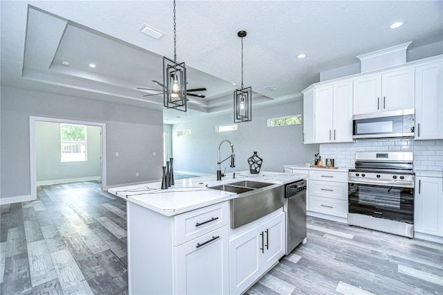 kitchen with hanging light fixtures, a raised ceiling, a center island with sink, white cabinets, and appliances with stainless steel finishes