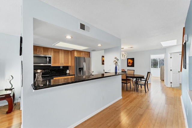 kitchen with kitchen peninsula, appliances with stainless steel finishes, light wood-type flooring, a skylight, and dark stone counters