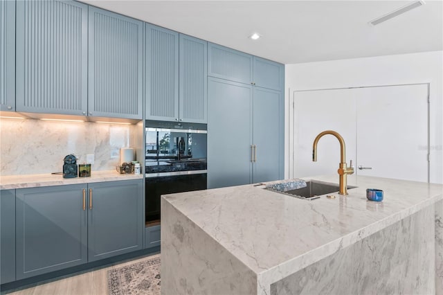 kitchen with a kitchen island with sink, sink, light hardwood / wood-style flooring, decorative backsplash, and light stone counters