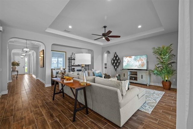 living room with a raised ceiling, ceiling fan with notable chandelier, dark hardwood / wood-style flooring, and ornamental molding