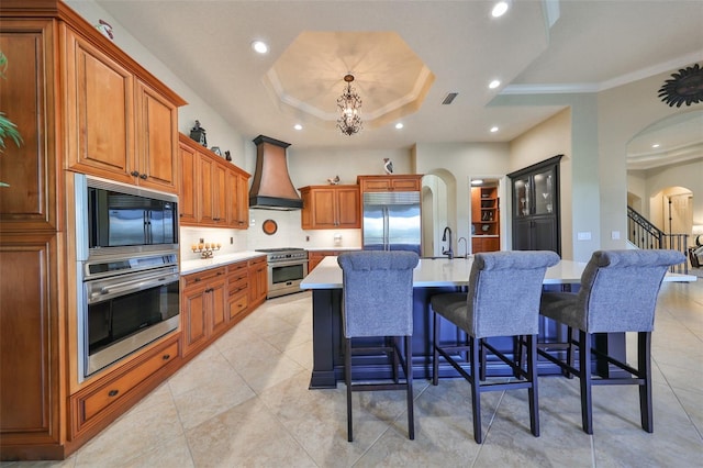 kitchen with custom exhaust hood, a kitchen bar, built in appliances, an island with sink, and a raised ceiling