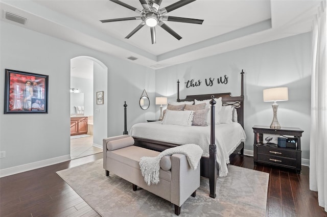 bedroom featuring a raised ceiling, ceiling fan, dark hardwood / wood-style flooring, and connected bathroom