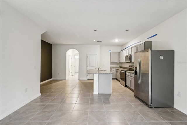 kitchen with light stone countertops, appliances with stainless steel finishes, gray cabinetry, a kitchen island with sink, and light tile patterned floors