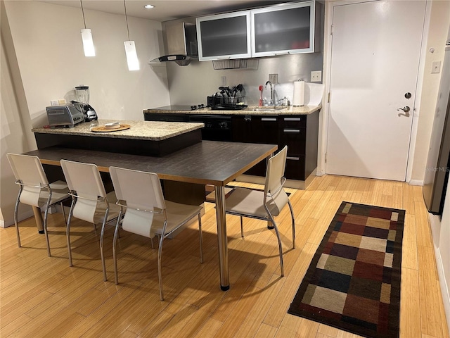 kitchen with sink, hanging light fixtures, wall chimney range hood, light stone counters, and light wood-type flooring