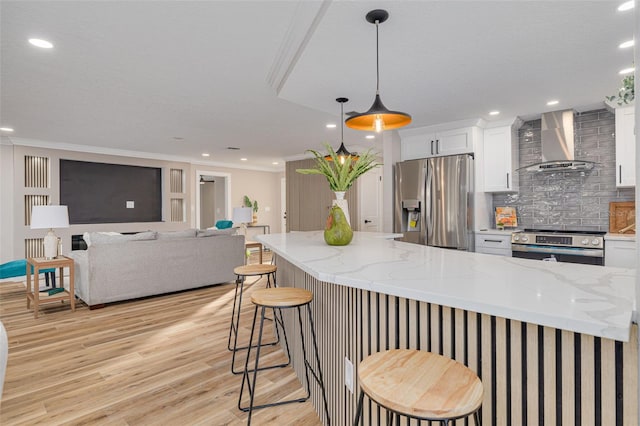 kitchen featuring wall chimney range hood, white cabinetry, hanging light fixtures, stainless steel appliances, and a kitchen bar