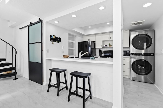 kitchen with white cabinetry, a kitchen breakfast bar, a barn door, stacked washer / drying machine, and black appliances