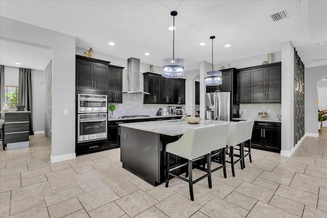 kitchen with decorative backsplash, appliances with stainless steel finishes, wall chimney exhaust hood, a kitchen island with sink, and decorative light fixtures