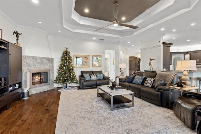 living room featuring a tray ceiling, ceiling fan, crown molding, dark wood-type flooring, and a premium fireplace