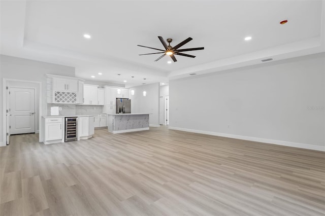 unfurnished living room with a tray ceiling, wine cooler, ceiling fan, and light hardwood / wood-style flooring