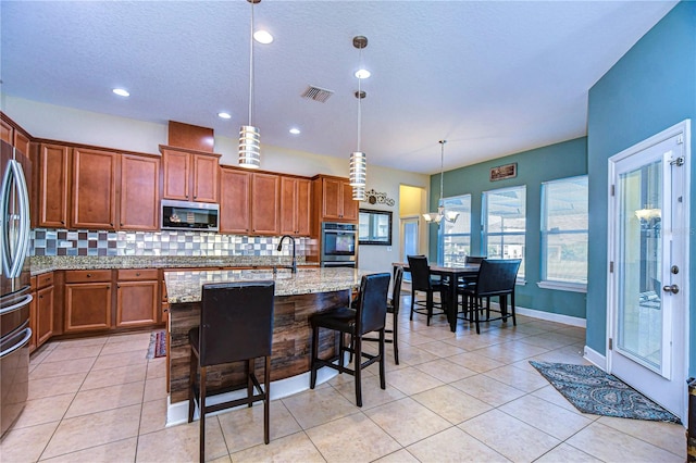 kitchen with light stone countertops, stainless steel appliances, an island with sink, decorative light fixtures, and a kitchen bar