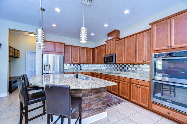 kitchen with black appliances, a center island with sink, sink, decorative light fixtures, and light stone counters