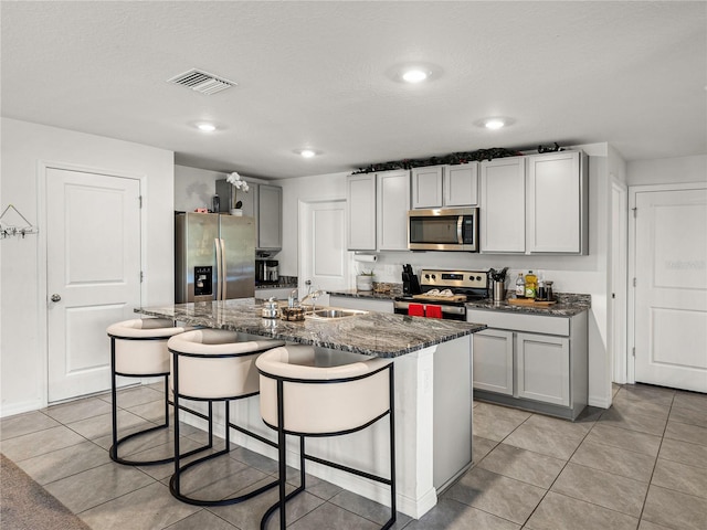 kitchen featuring sink, stainless steel appliances, dark stone counters, gray cabinets, and a kitchen island with sink