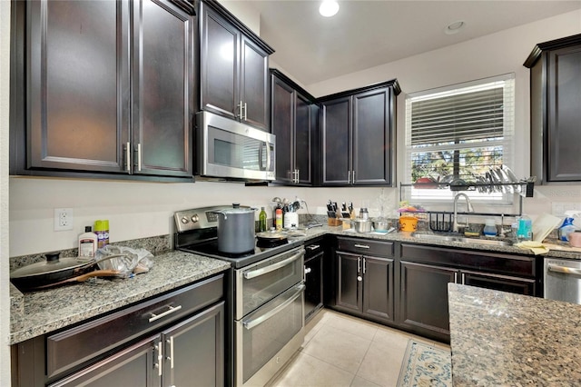 kitchen with appliances with stainless steel finishes, light stone counters, dark brown cabinetry, sink, and light tile patterned floors