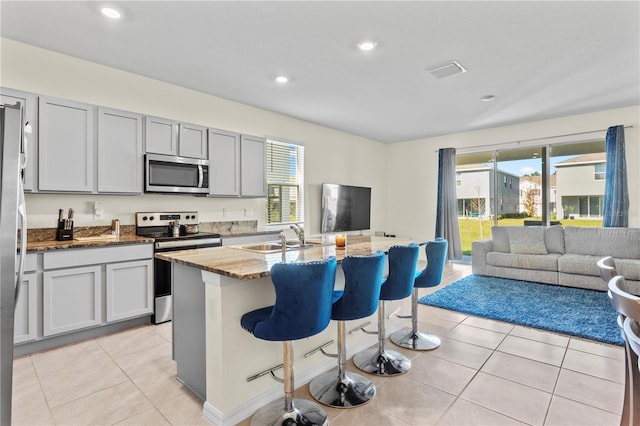 kitchen with sink, appliances with stainless steel finishes, a kitchen island with sink, light stone counters, and a kitchen bar