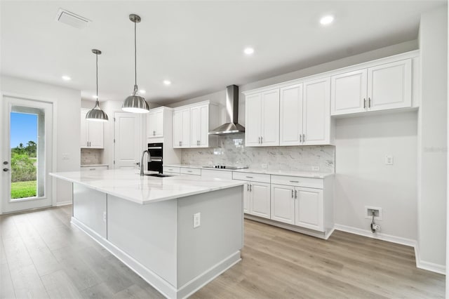 kitchen featuring light stone countertops, wall chimney exhaust hood, light hardwood / wood-style flooring, a kitchen island with sink, and white cabinets