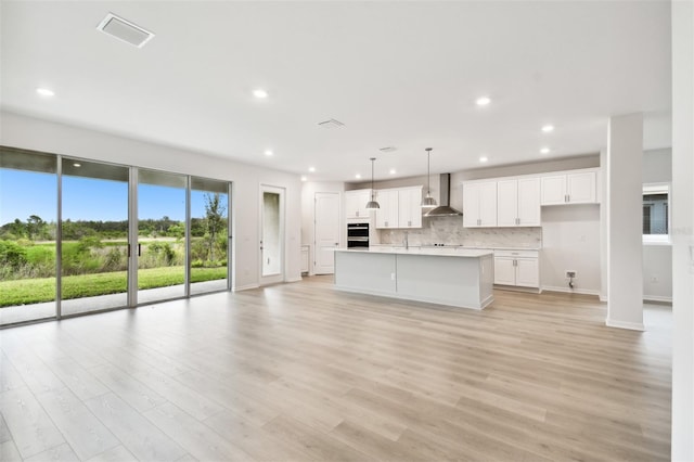 kitchen featuring light wood-type flooring, wall chimney range hood, pendant lighting, white cabinets, and an island with sink