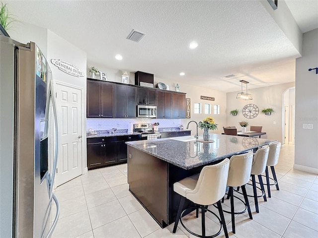 kitchen featuring pendant lighting, a center island with sink, a textured ceiling, appliances with stainless steel finishes, and dark brown cabinetry