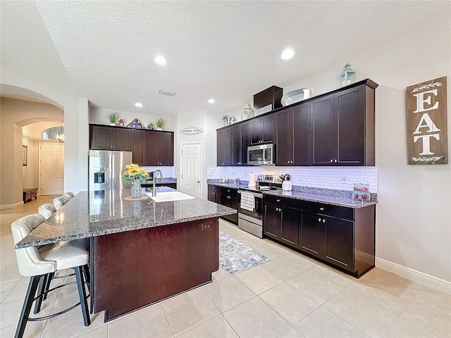 kitchen featuring a center island with sink, light stone countertops, dark brown cabinetry, and appliances with stainless steel finishes