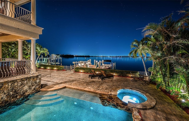 pool at night featuring a water view, an in ground hot tub, a patio, and a dock