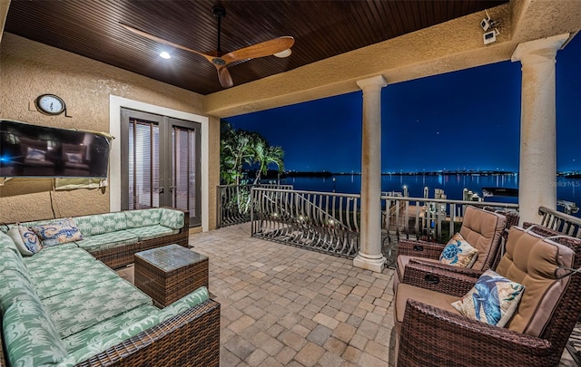 patio at night with ceiling fan, a water view, an outdoor hangout area, and a balcony