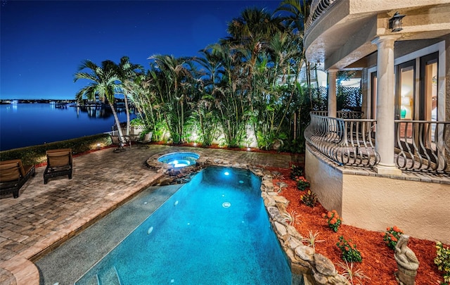 pool at night with an in ground hot tub, a water view, and a patio