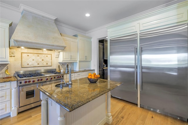 kitchen featuring high quality appliances, premium range hood, a center island with sink, sink, and decorative backsplash