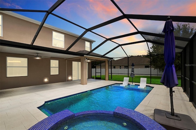 pool at dusk featuring glass enclosure, ceiling fan, a yard, an in ground hot tub, and a patio