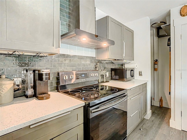 kitchen with appliances with stainless steel finishes, backsplash, gray cabinetry, wall chimney range hood, and light hardwood / wood-style floors