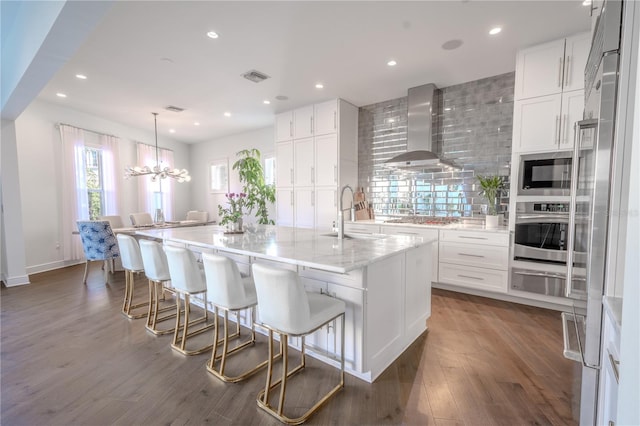 kitchen featuring sink, a large island with sink, appliances with stainless steel finishes, wall chimney range hood, and white cabinets