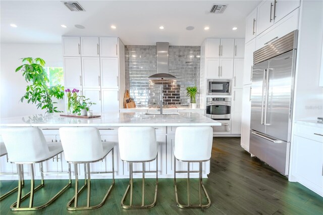 kitchen with white cabinetry, a breakfast bar area, wall chimney range hood, built in appliances, and a center island with sink