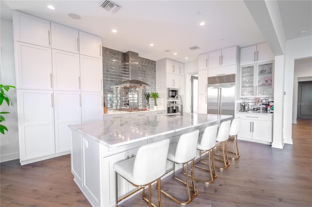 kitchen featuring a spacious island, wall chimney exhaust hood, light stone counters, and white cabinets