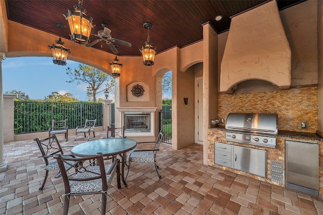 patio terrace at dusk featuring ceiling fan, a grill, exterior kitchen, and an outdoor fireplace