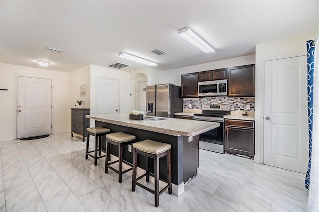 kitchen featuring a kitchen island with sink, decorative backsplash, dark brown cabinets, a kitchen bar, and stainless steel appliances