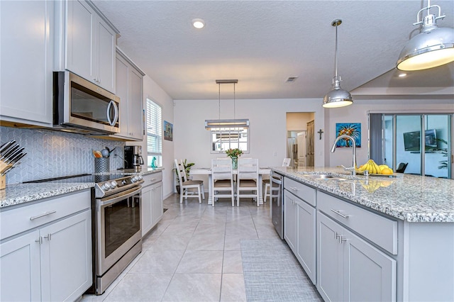 kitchen with appliances with stainless steel finishes, tasteful backsplash, gray cabinetry, a kitchen island with sink, and decorative light fixtures