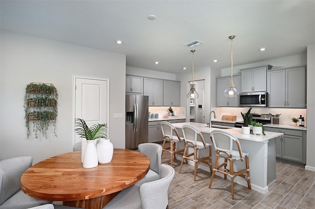 kitchen with sink, gray cabinets, an island with sink, appliances with stainless steel finishes, and decorative light fixtures