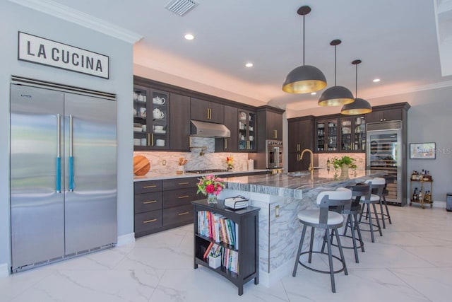 kitchen with dark brown cabinetry, hanging light fixtures, dark stone countertops, a kitchen island with sink, and appliances with stainless steel finishes