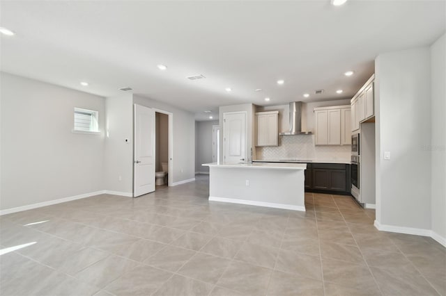 kitchen with wall chimney range hood, light tile patterned floors, appliances with stainless steel finishes, a kitchen island with sink, and tasteful backsplash