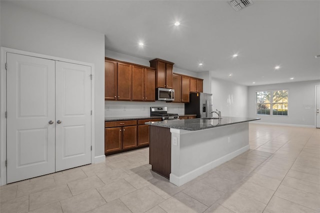 kitchen featuring a kitchen island with sink, dark stone counters, sink, light tile patterned floors, and appliances with stainless steel finishes